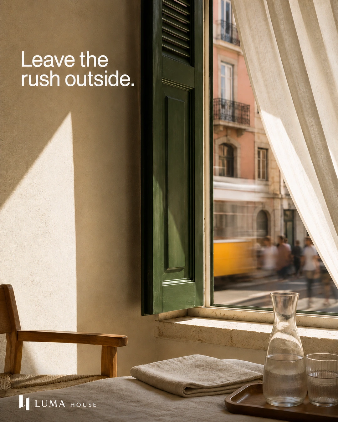 A still sunlit room with linen and a carafe contrasts with a blurred tram and pedestrians beyond a green shutter.